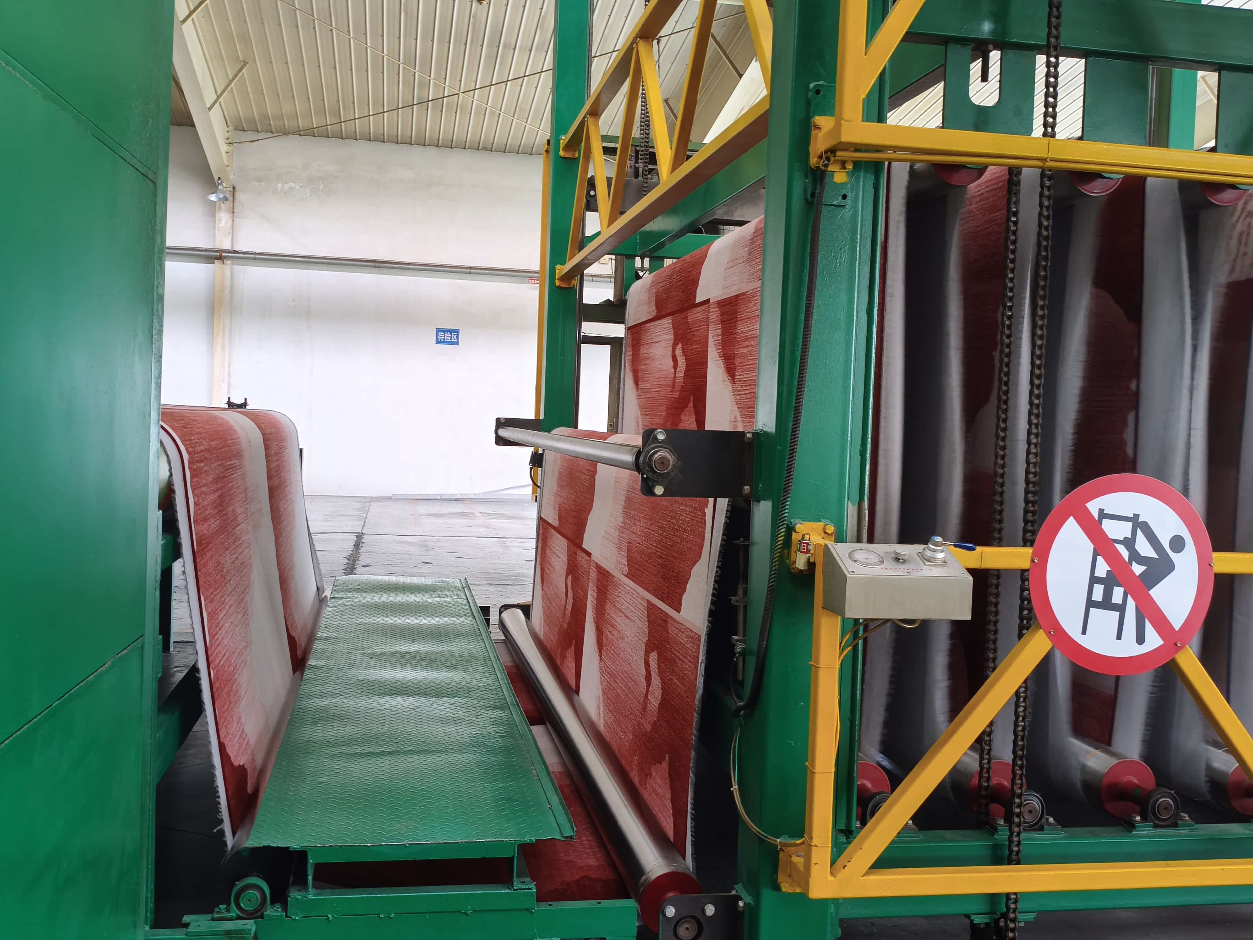 Printed carpet roll in red and white being fed into automated finishing machine with safety barriers