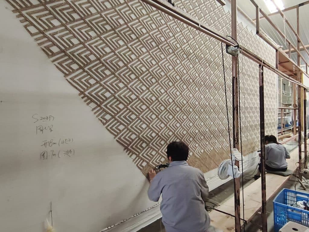 Hand tufted carpet workshop with workers tufting large geometric pattern in brown on stretched backing cloth