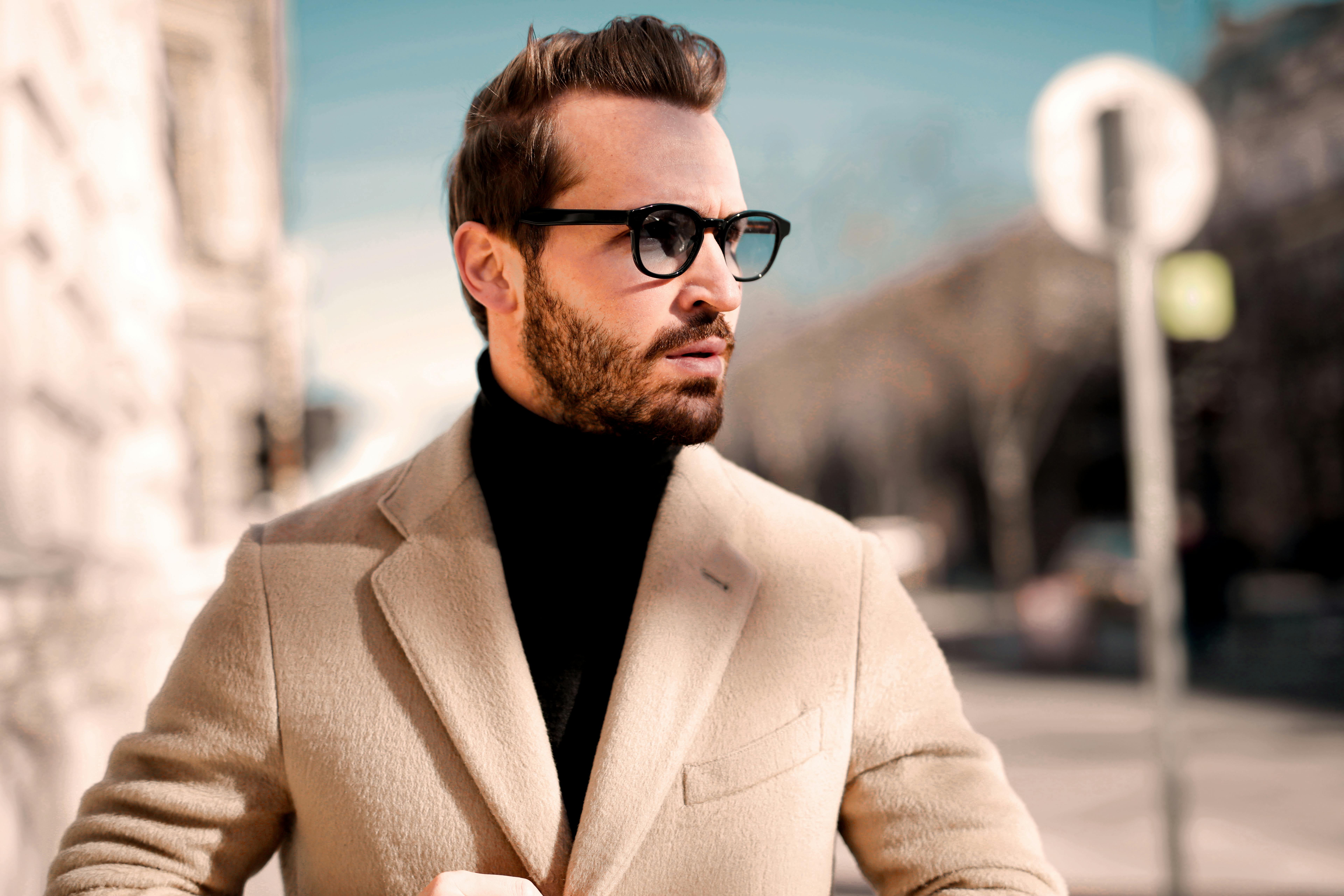 Stylish man in a beige coat and sunglasses looking away on a sunny city street.